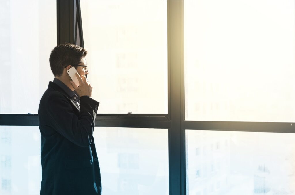 **Titel:** Geschäftsmann bei der Telefonkommunikation vor großen Fenstern **Alt-Beschreibung:** Ein Geschäftsmann in einem Anzug telefoniert mit seinem Smartphone und blickt dabei durch große Bürofenster in helles Licht.